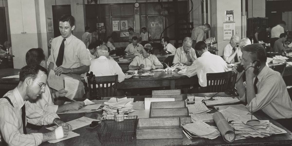 White journalists sit around a busy news room, black and white